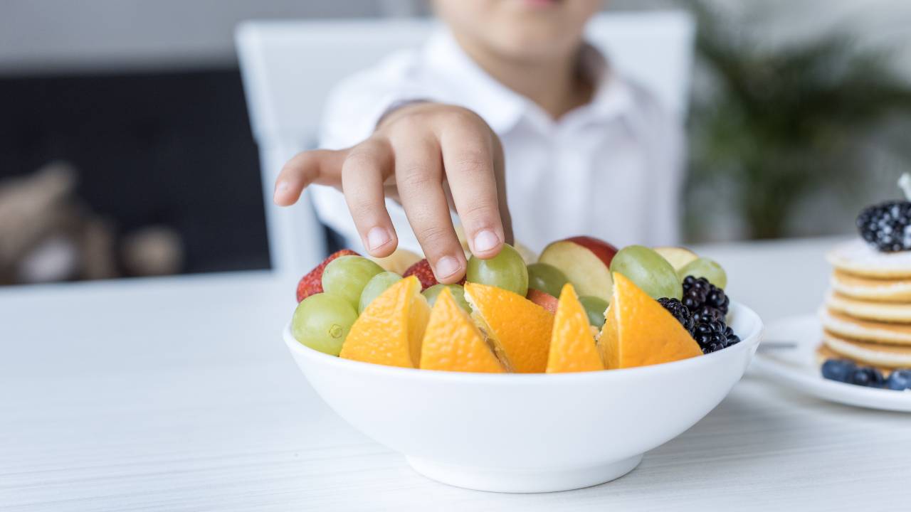 Criança pegando fruta de tigela com diversas frutas.