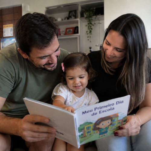 Família lê um livro sobre escola com a filha de 2 anos, preparando para a adaptação escolar com confiança e alegria.